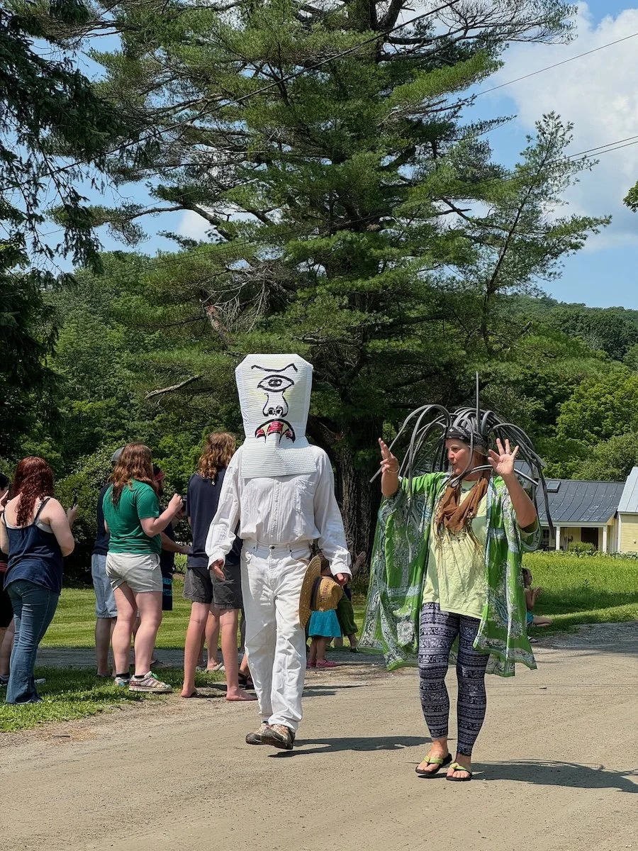 People dressed in costumes, one wearing a large white mask with a painted face, and the other with a costume that has long black wires or tentacles, walking outdoors on a dirt path with trees and a house in the background.