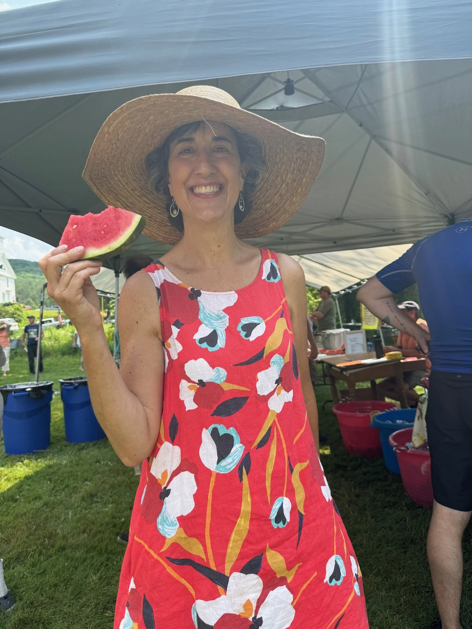 A woman wearing a large straw hat and a red floral dress, smiling at the camera, holding a slice of watermelon outdoors under a tent with other people and tents in the background.