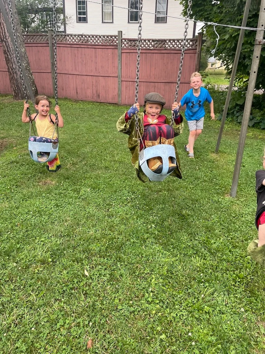 Three children playing on a swing set in a backyard, with two girls on swings and a boy standing nearby, all smiling.