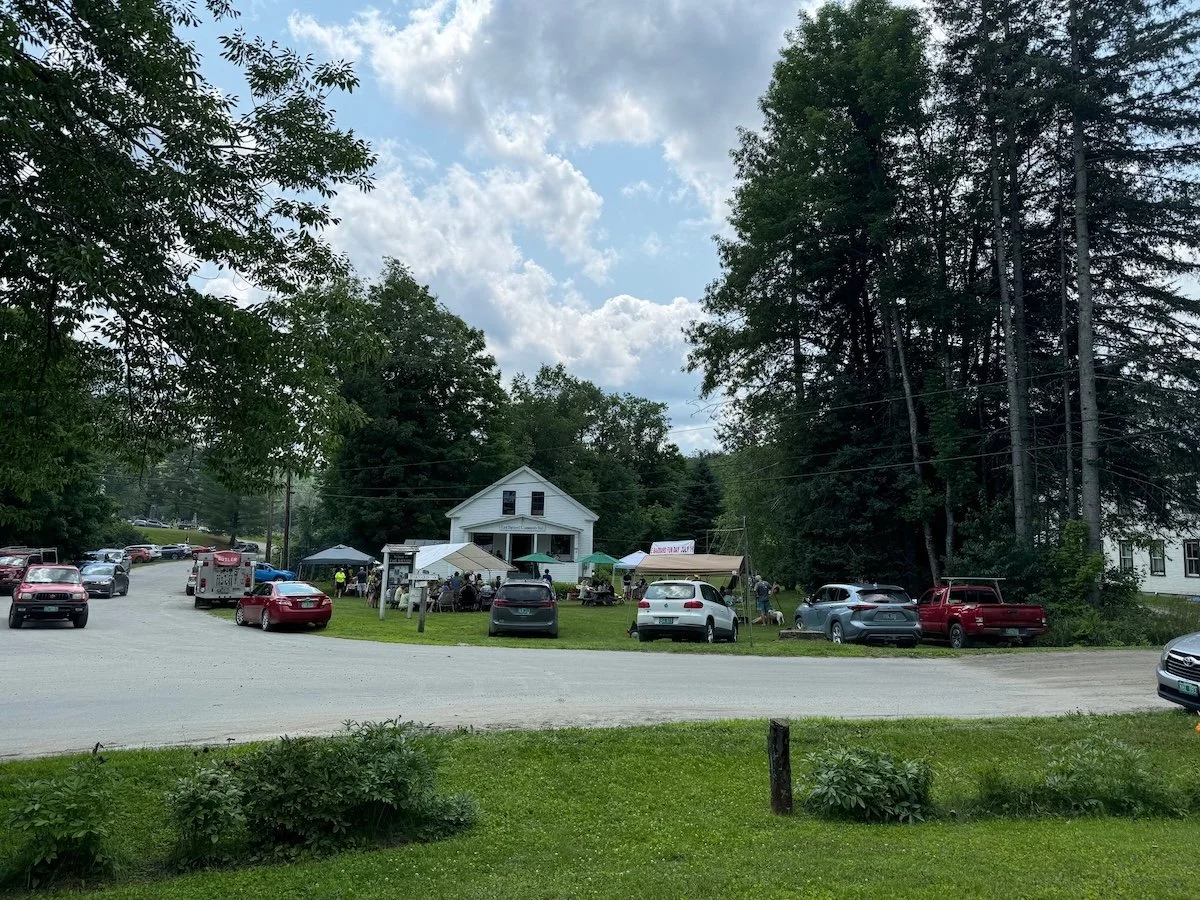 A rural outdoor event with several tents and a crowd gathered in front of a white building, surrounded by trees and parked cars, under a partly cloudy sky.