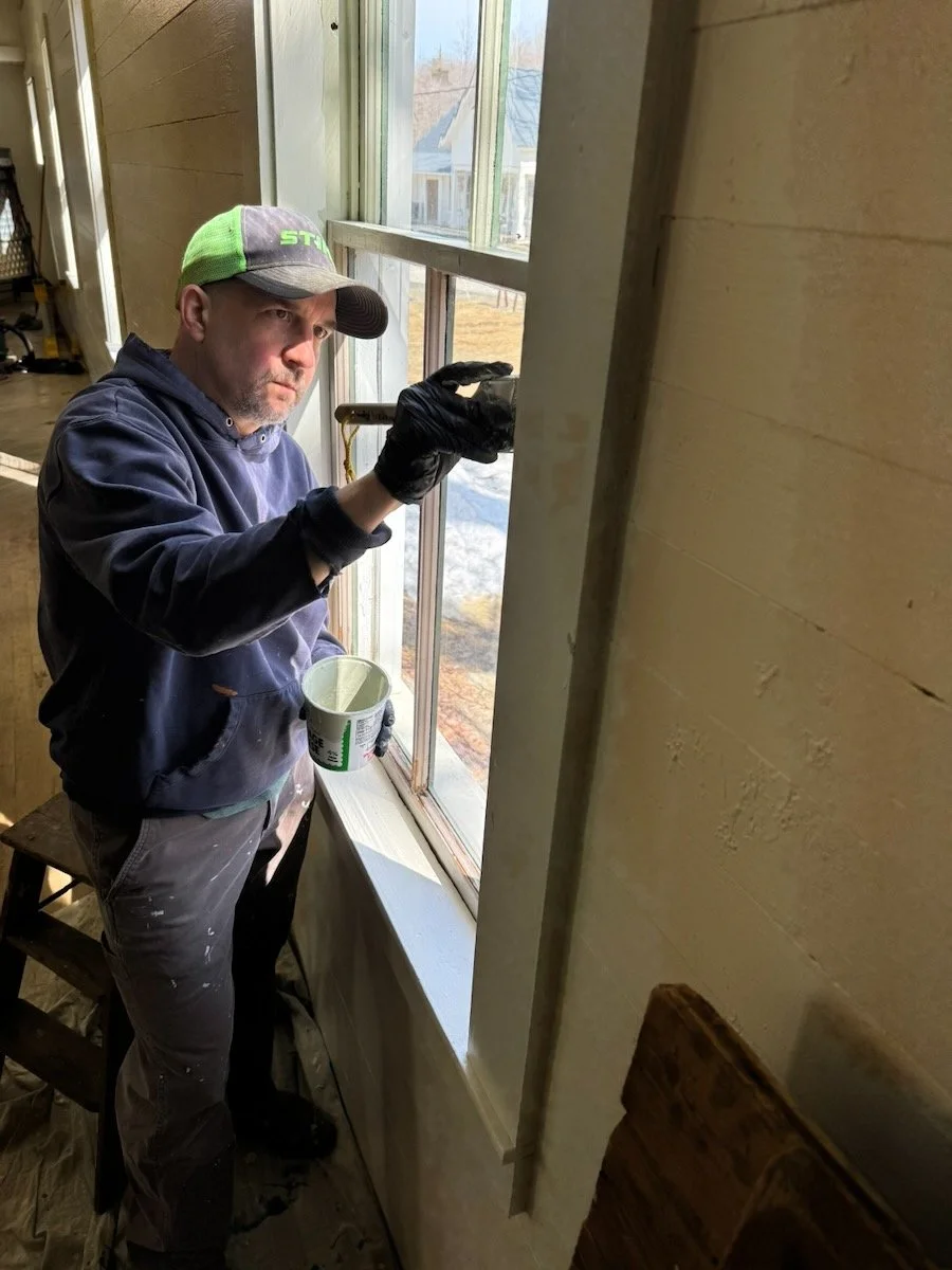 A man wearing a green and gray cap, black gloves, and dark clothing is applying paint to a window frame with a brush, holding a small container of paint, in a room with wood paneling on the walls and a view of an outdoor area through the window.