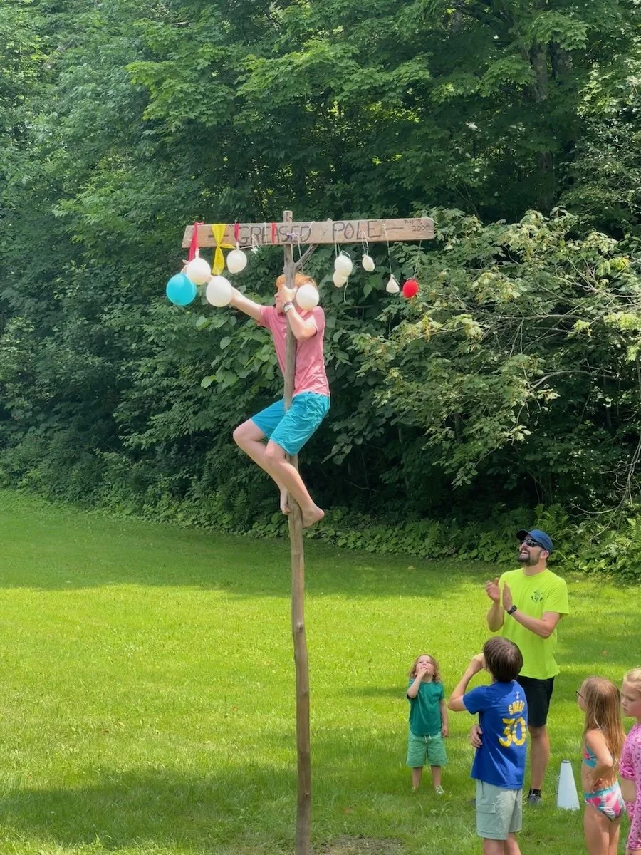 A person climbing a tall wooden pole to reach a horizontal wooden bar decorated with balloons, with a sign reading 'GREAT POLE', as children and an adult watch and applaud outdoors on a grassy field surrounded by trees.