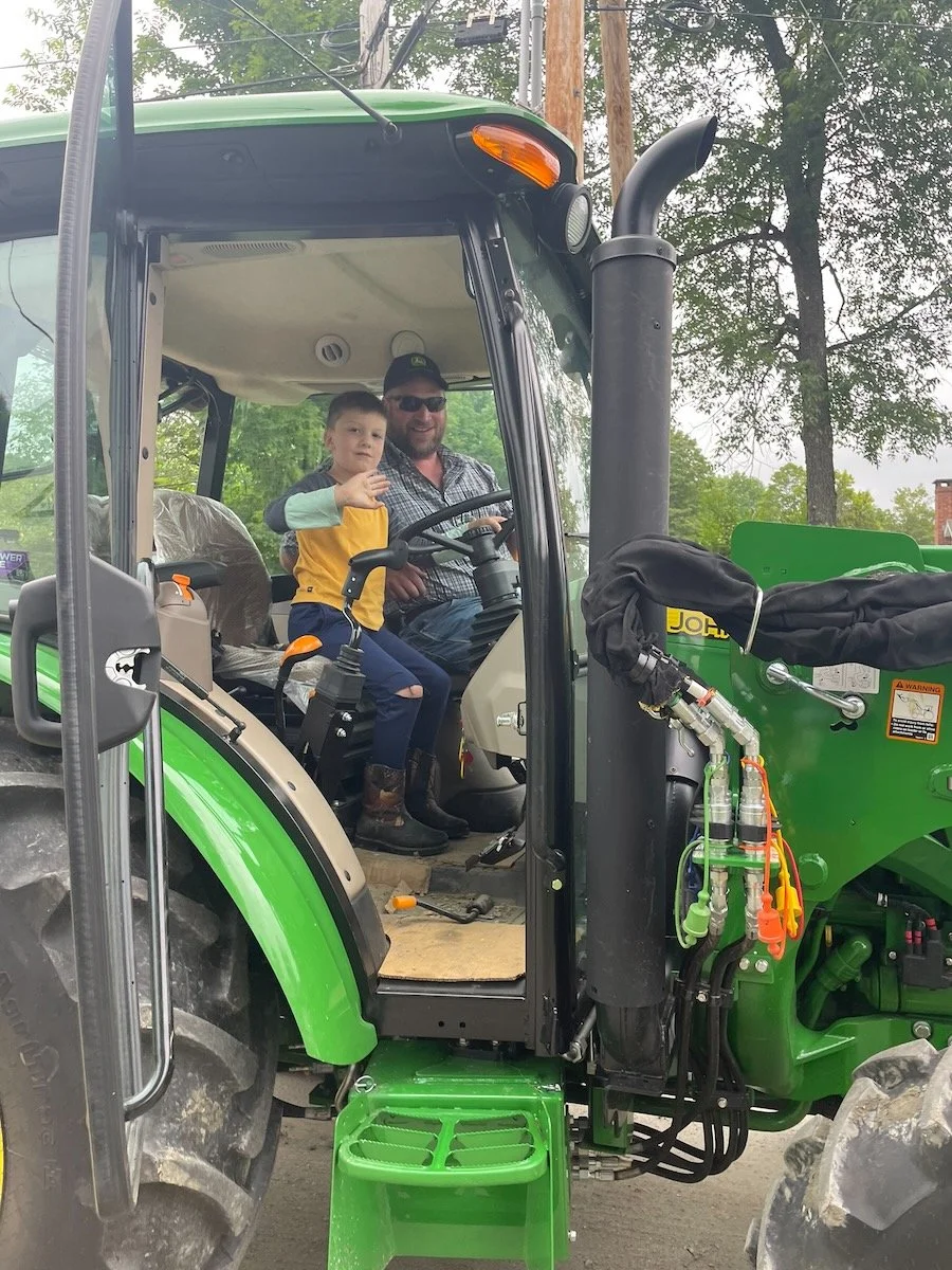 A man and a young boy sitting inside the cabin of a green John Deere tractor, smiling and waving at the camera. The background shows trees and power lines.