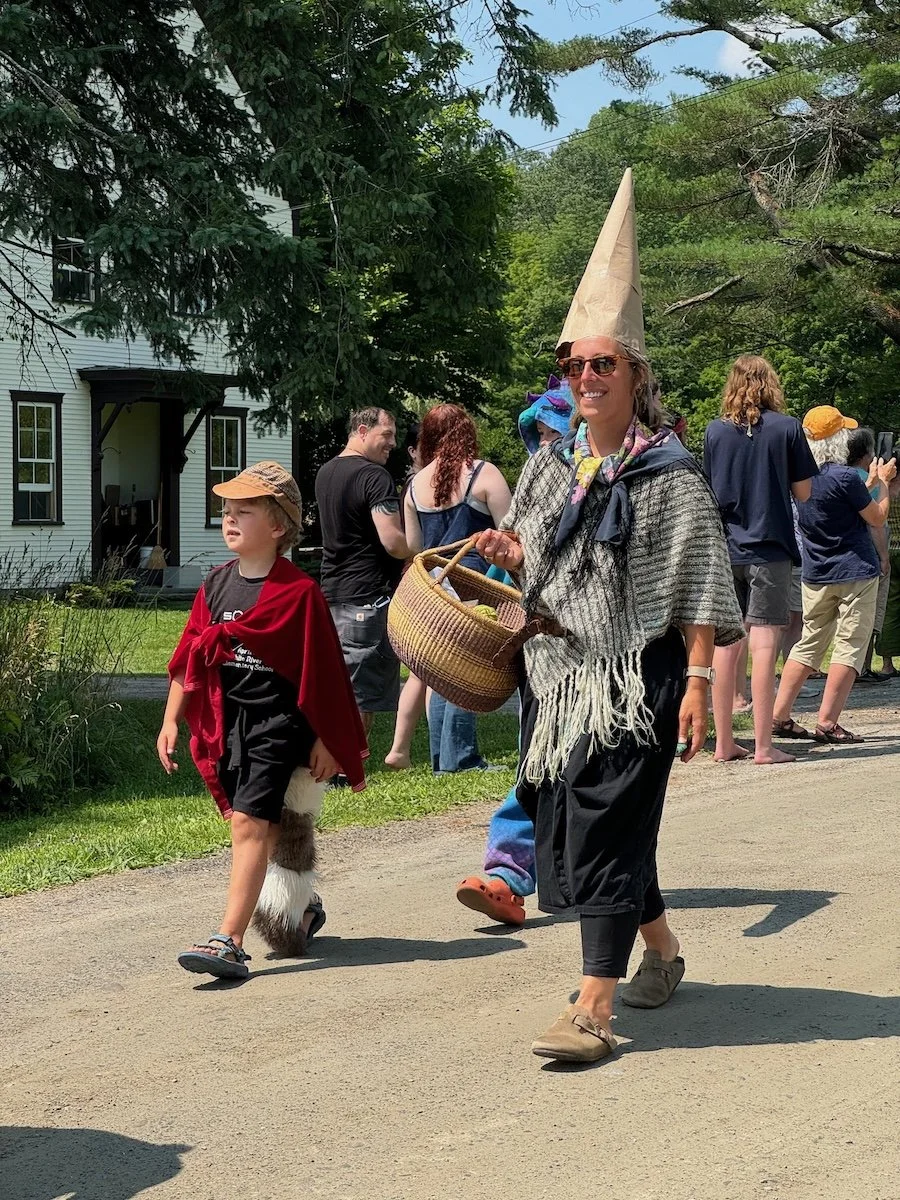 A woman wearing a tall paper cone hat and sunglasses, smiling, walking outdoors with a basket, surrounded by people in casual and costume clothing during a parade or festival on a sunny day.