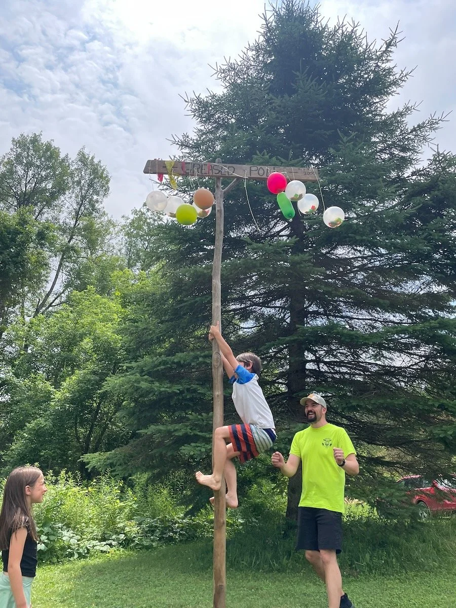 A person climbing a tall wooden pole with balloons attached at the top, labeled 'Tie-Dye Pokes,' while two children watch and laugh outdoors with trees in the background.