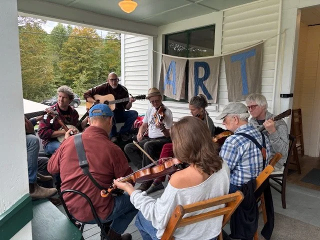 A group of people playing instruments together on a porch outdoors, with a banner that says "ART" hanging behind them.