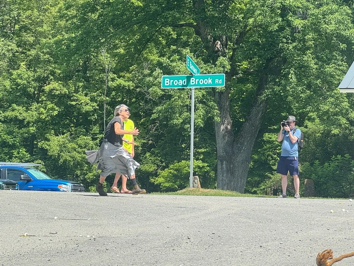 A woman with gray hair running, with a person in a yellow vest and another in a pink shirt behind her, near a street sign at Broad Brook Road and East Street. A man in a cap is taking a photo on the right side of the image. There are trees and parked