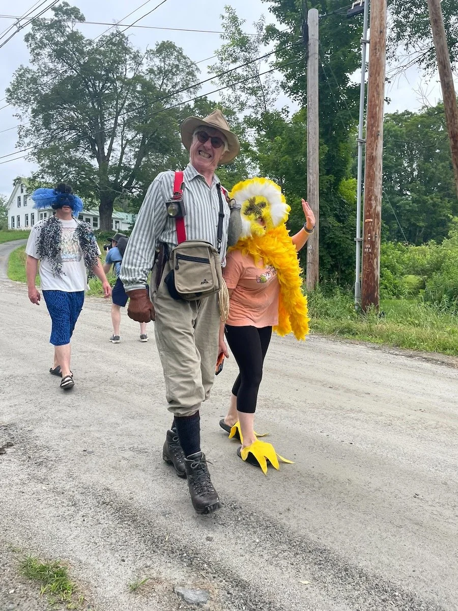 A group of people walking outdoors on a gravel road, some dressed in colorful and humorous costumes, including a person in a chicken costume and another in a feathered headdress, with trees, utility poles, and a house visible in the background.
