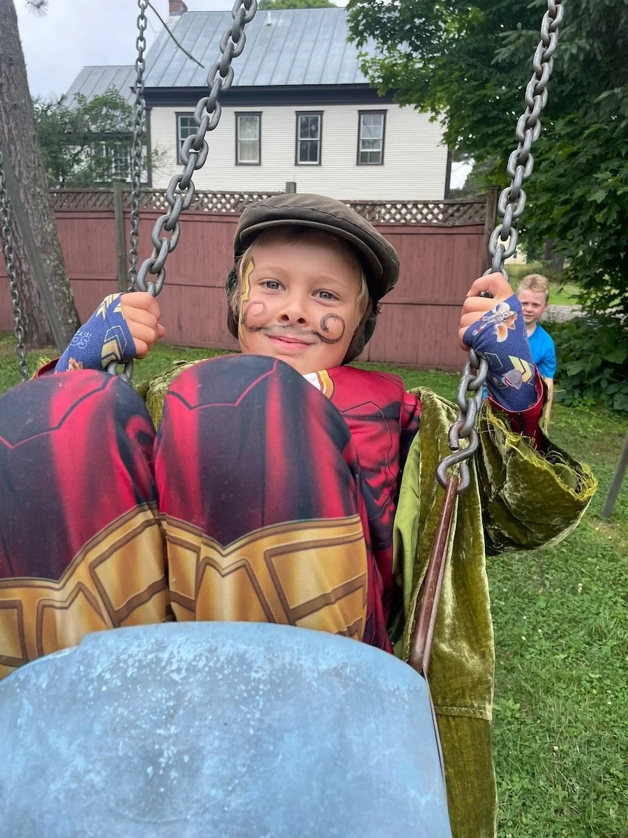 A young boy with face paint, dressed in a superhero costume, sitting on a swing outdoors in a backyard. Another child is visible in the background.
