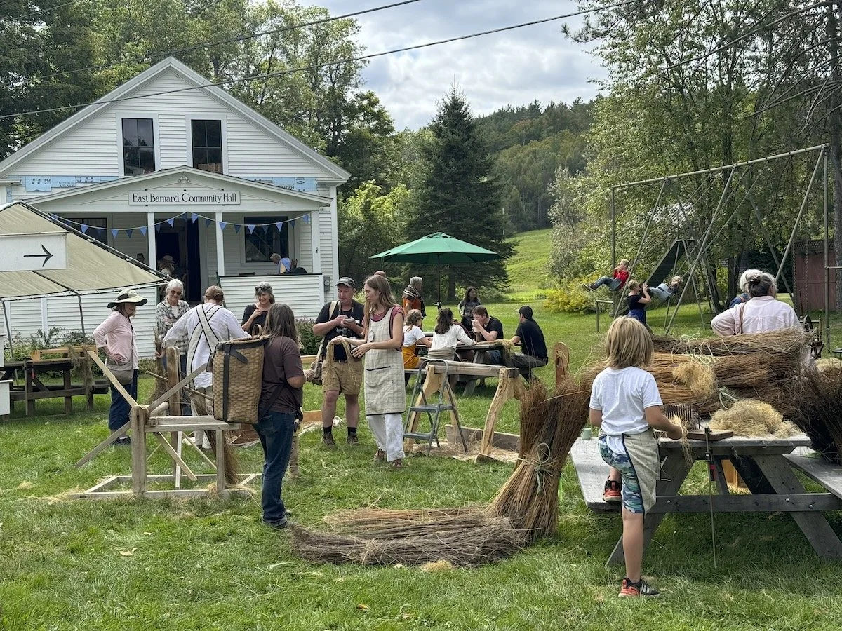 Community gathering outside the East Barnard Community Hall with people socializing, children playing on swings, and a woman preparing traditional broom-making materials on a workbench.