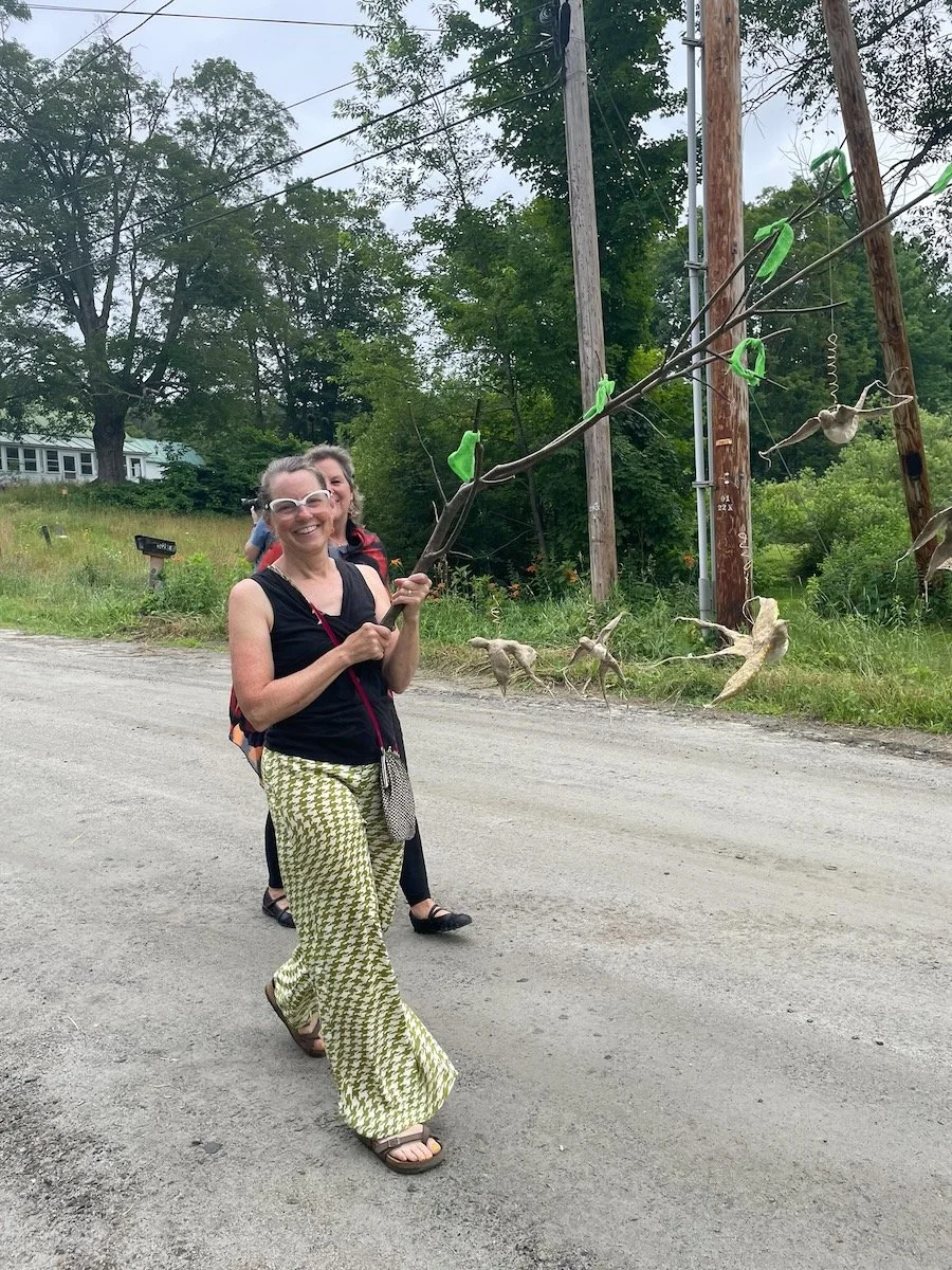 Woman standing outdoors on a dirt road, holding a large stick with hanging cloth and paper decorations, smiling at the camera. Background shows trees and utility poles.