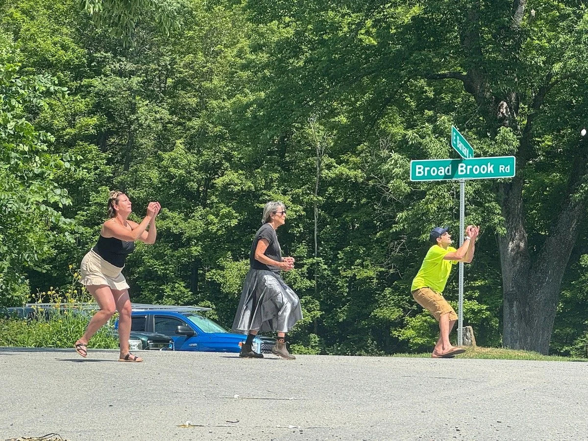 Three people are standing on the side of the road near a street sign that reads "Broad Brook Rd." The person on the right, wearing a yellow shirt and shorts, is holding a camera or phone, aiming it upward. The middle person is wearing a black t-shirt