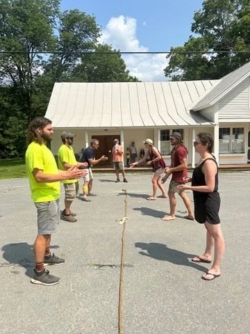 People playing a game with a rope outdoors near a building with a metal roof and trees in the background.