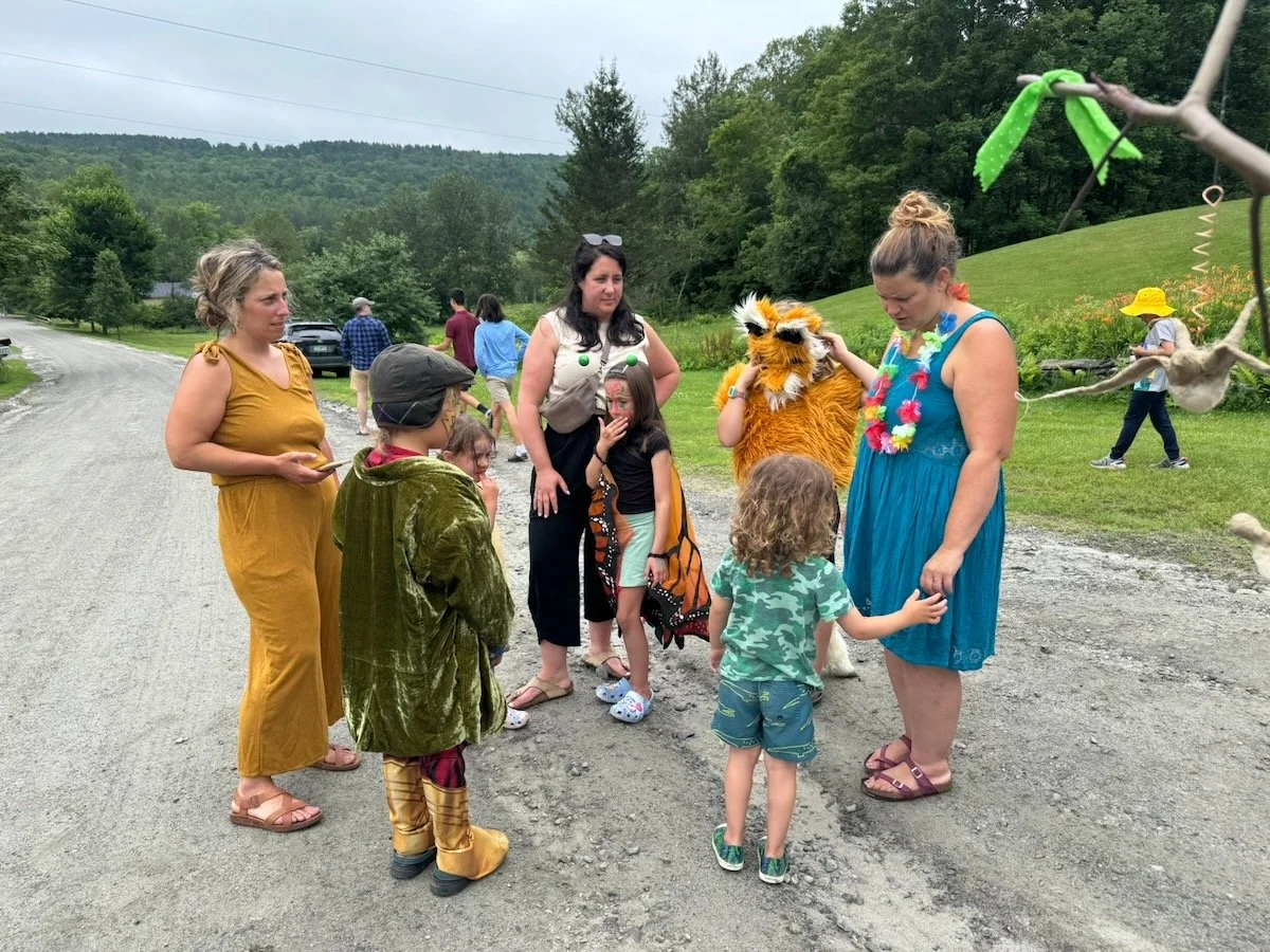 People dressed in costumes, including animals and jungle-themed outfits, gathered outdoors on a gravel road surrounded by green trees and hills, participating in a festive event.
