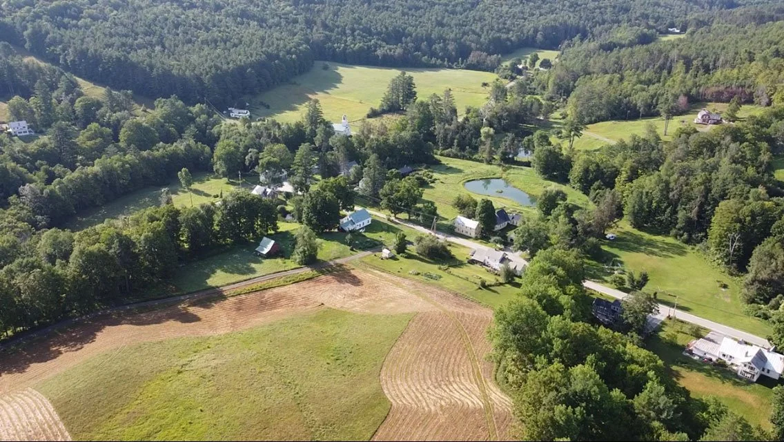 Aerial view of a rural landscape with trees, houses, ponds, fields, and a winding road.