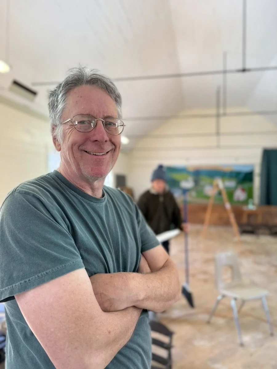 A smiling middle-aged man with gray hair, glasses, wearing a gray t-shirt, standing with arms crossed in a room with a blurred person in the background, a chair, and a colorful mural.