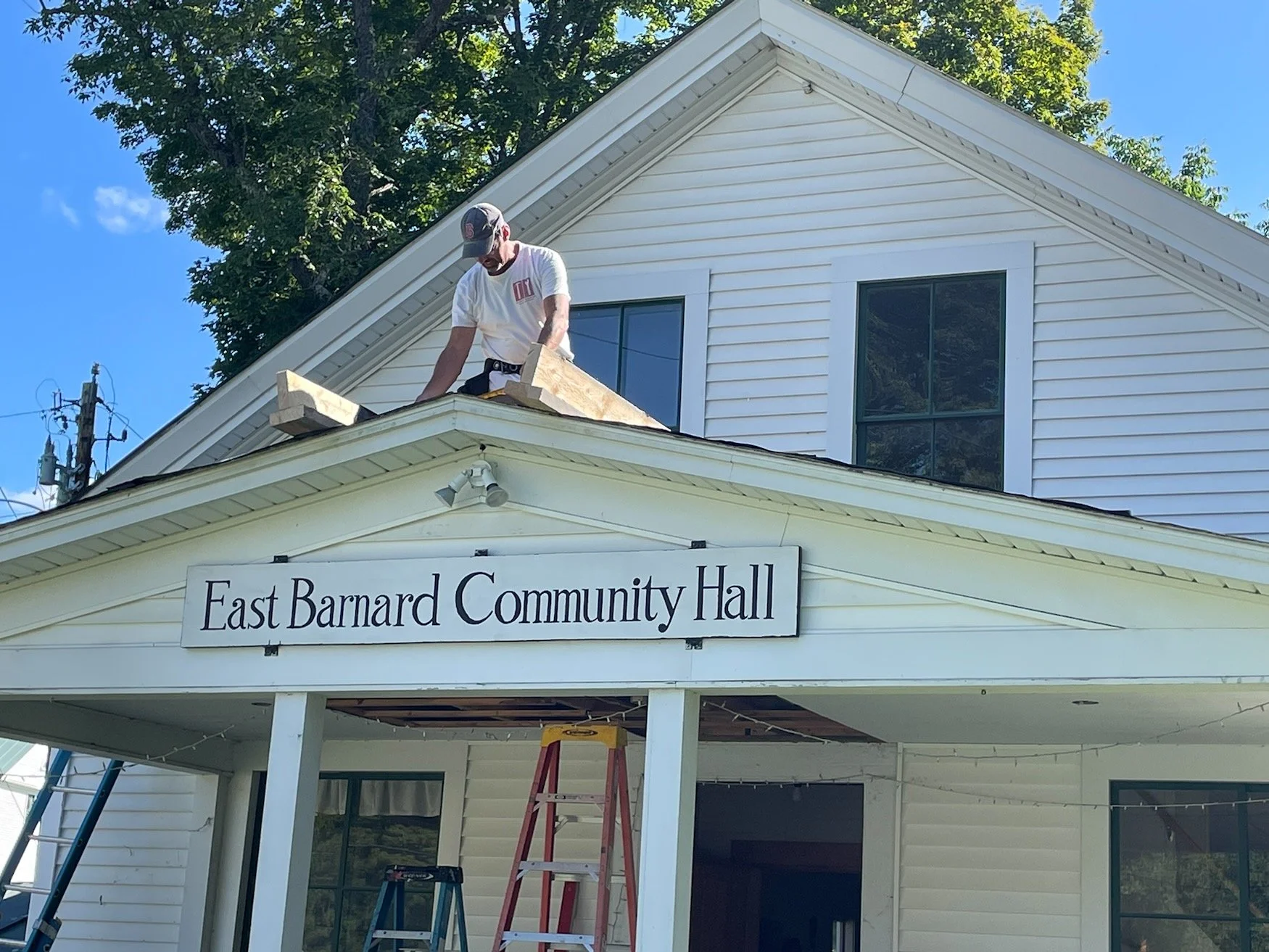People working on the roof of the East Barnard Community Hall building on a ladder, with trees and a blue sky in the background.