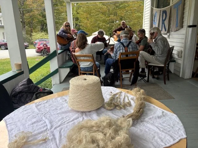 A group of people gathered on a porch, playing guitars and singing together during the daytime, with an outdoor setting in the background. In the foreground, a table with a large spool of yarn and some loose yarns.