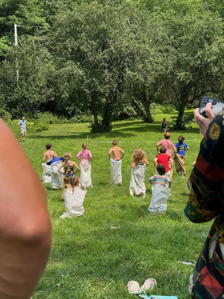 Children playing a sack race in a grassy outdoor area with trees, while adults watch and photograph.