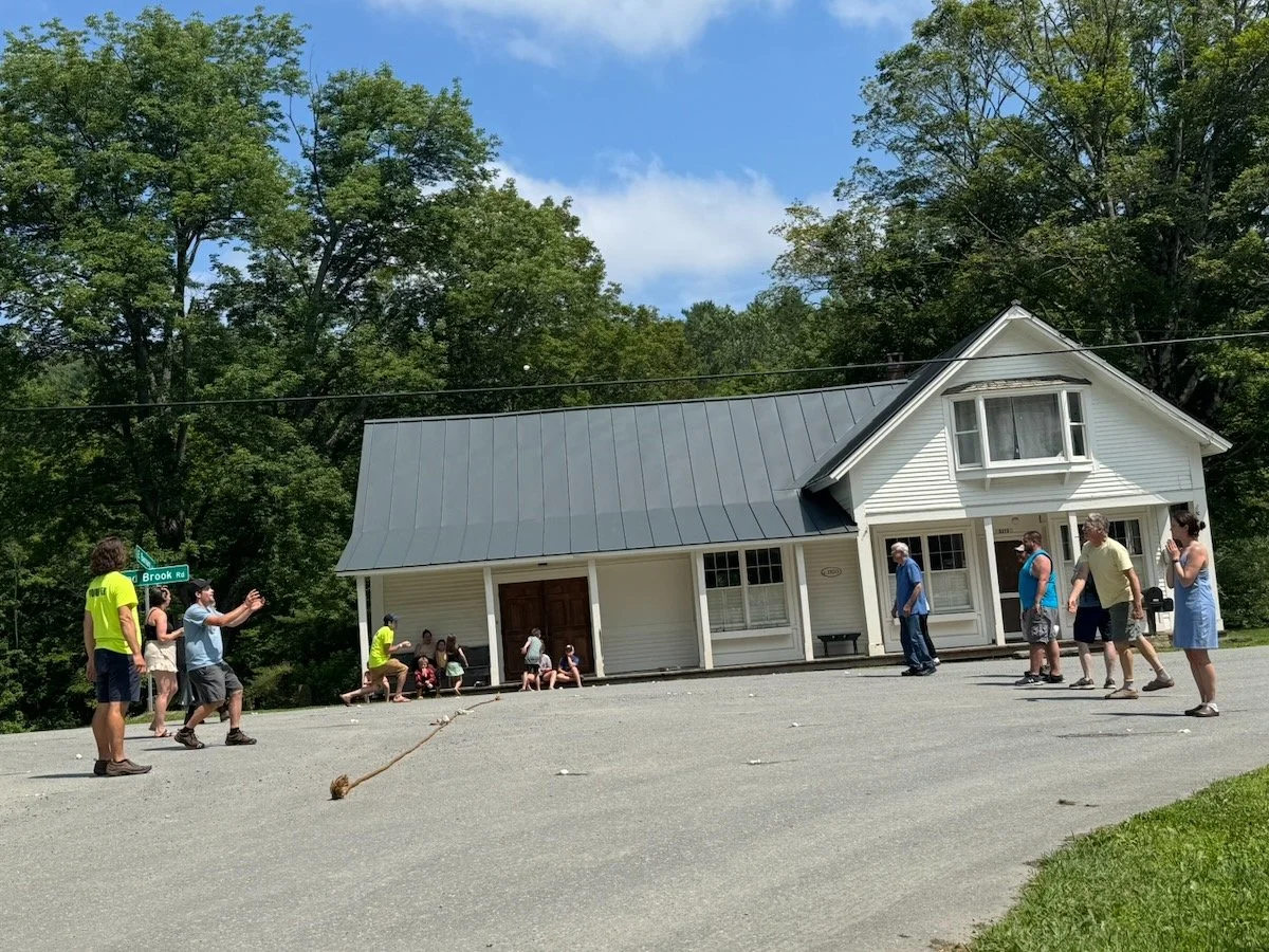 People gathered outside a white house with a gray metal roof, playing a game with a rope in the driveway on a sunny day, surrounded by trees.