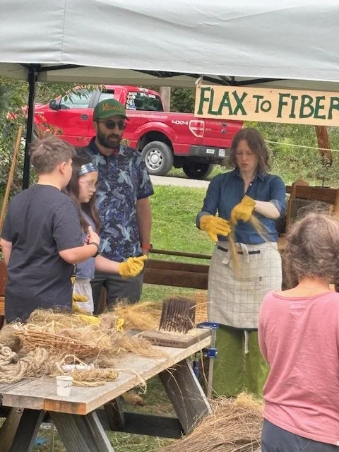 People at a booth demonstrating flax to fiber process, with a sign reading 'Flax to Fiber', and a red truck in the background.