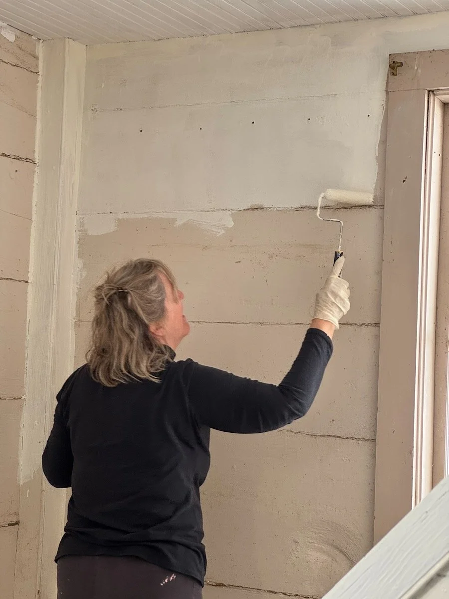 A woman painting a wall with a roller brush, wearing gloves and a black shirt.