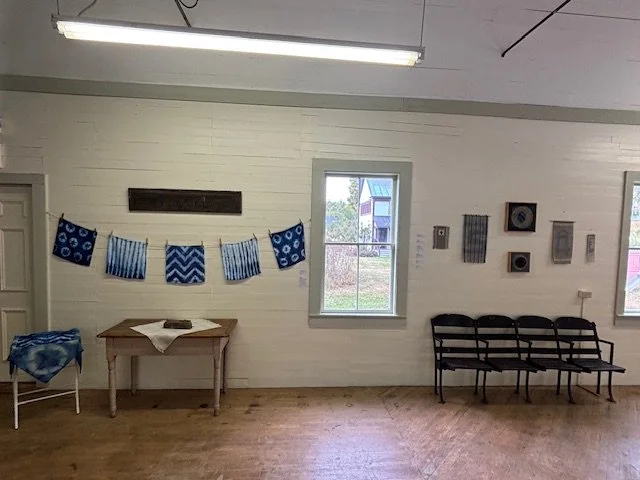 An interior room with white wooden walls, two windows showing a grassy area and a house outside, a row of six black chairs, a wooden table with a blue and white cloth cover and a book or cloth on top, blue and white decorative fabrics hanging on the 