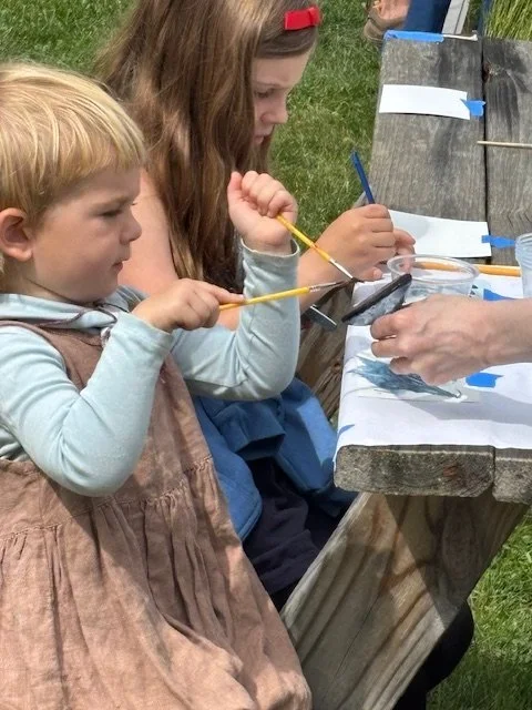 Two young girls sitting at a picnic table outdoors, participating in a craft activity with an adult assisting them.