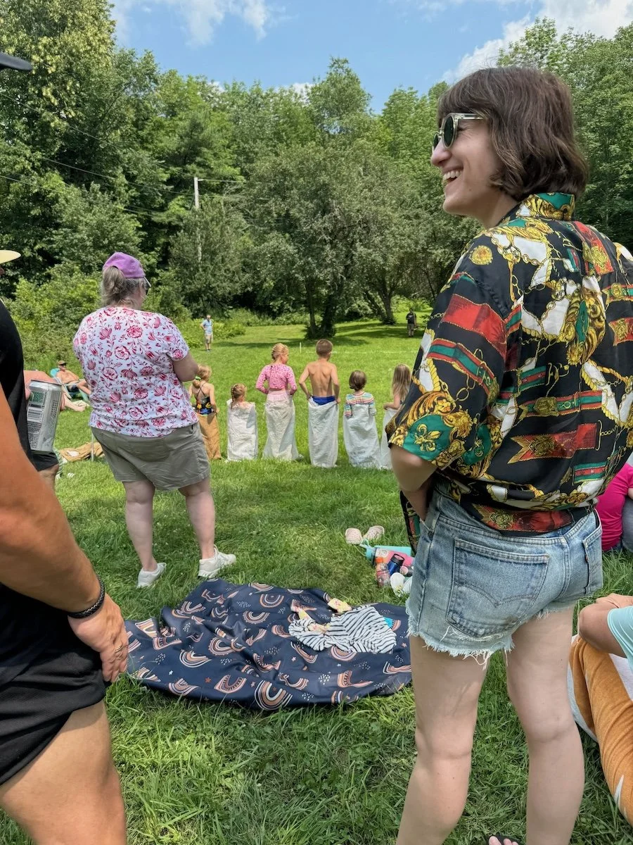 People outdoors watching a children's event, with some sitting on the grass and others standing, including a woman wearing sunglasses, a short-sleeved shirt with a colorful pattern, and denim shorts.