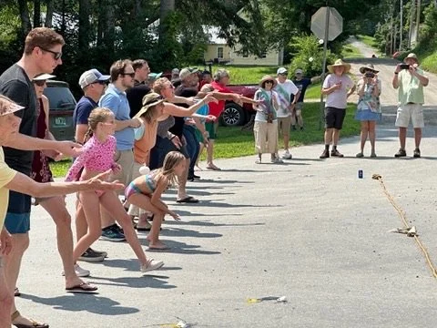 Group of people, including children and adults, participating in an outdoor game on a sunny day. They are lined up, stretching their arms forward, likely playing a game of tug-of-war or a similar activity, with trees, cars, and houses visible in the 