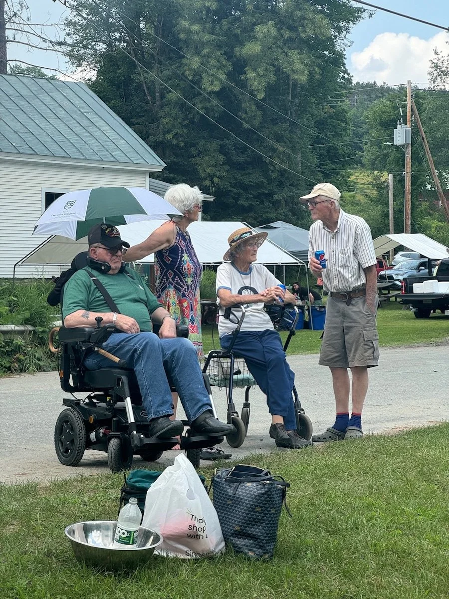 Four elderly people talking outdoors at a community gathering, one woman holding a beer, a man with a beverage, two women with mobility aids, with picnic items on the grass.