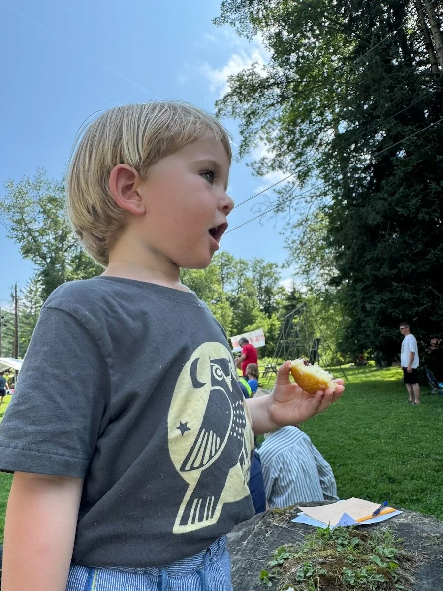 Young boy with blond hair wearing a dark t-shirt with a graphic design, holding a half-eaten corn dog, outdoors on a grassy area with trees, blue sky, and other people in the background.