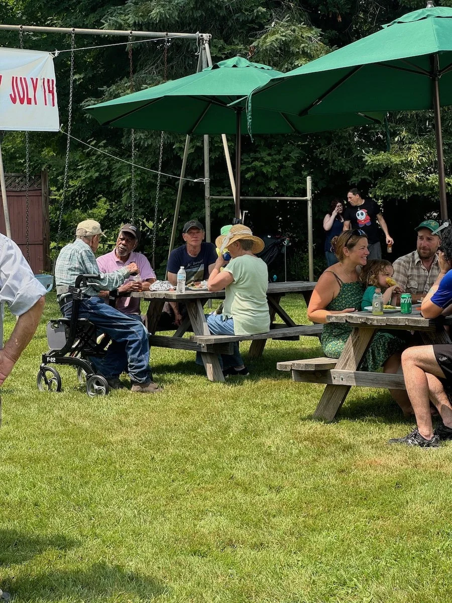 People gathered outdoors at picnic tables with green umbrellas, enjoying food and drinks, in a backyard setting with grassy lawn and trees in the background.
