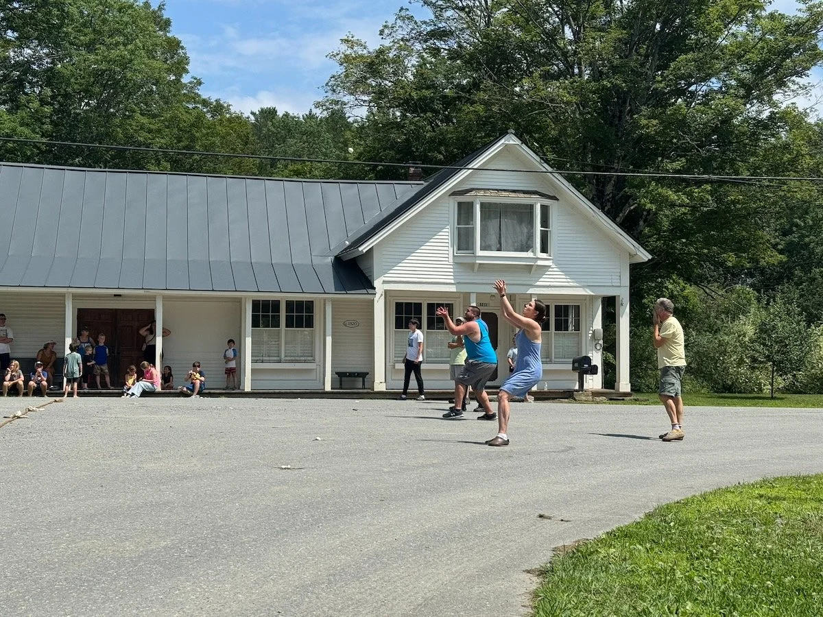 People playing a game of Tackle or Dodge outside a white house with a porch, surrounded by trees and grass on a sunny day.