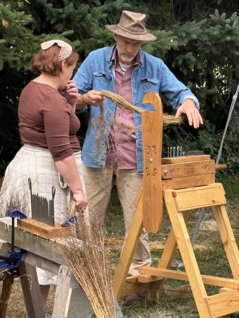 Two people, a woman and a man, are outdoors using a traditional wooden flax-spinning wheel to turn flax fibers into thread or yarn. The woman is watching attentively as the man operates the wheel, pulling fibers from a bundle.