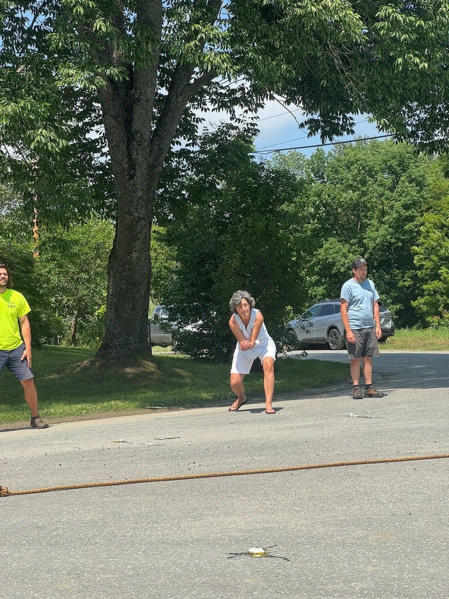 Three people standing outdoors on a sunny day, with large trees and parked cars in the background. One woman in a white sleeveless dress is bending down, while two men, one in a neon yellow shirt and another in a light blue shirt, stand nearby.