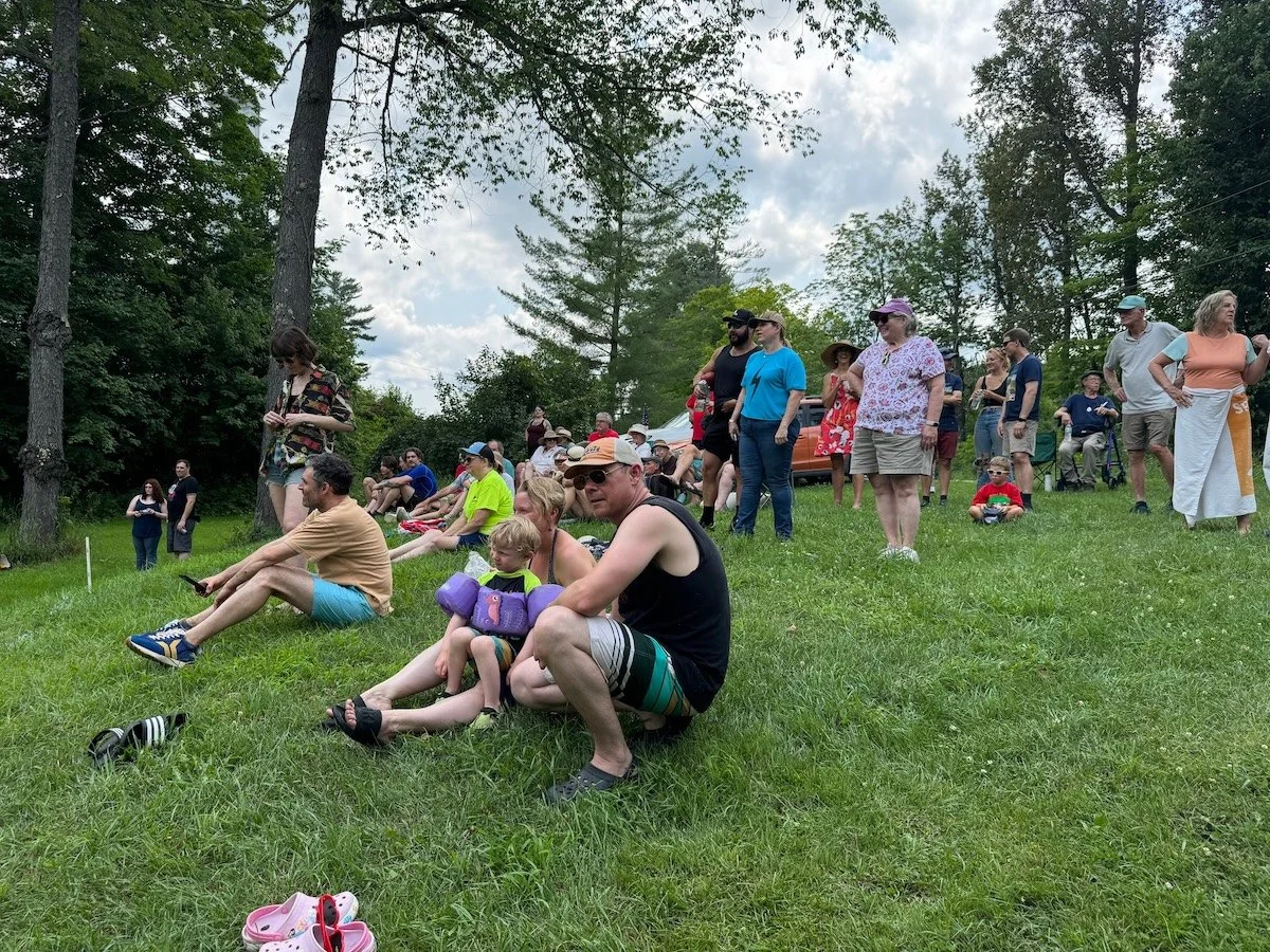 People gathered outdoors on a grassy hill under trees, some sitting and some standing, watching an event in the daytime.