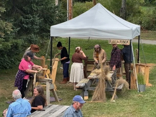 People demonstrating traditional fiber arts at a craft fair under a white canopy tent, with a sign that reads 'Flax to Fiber.'