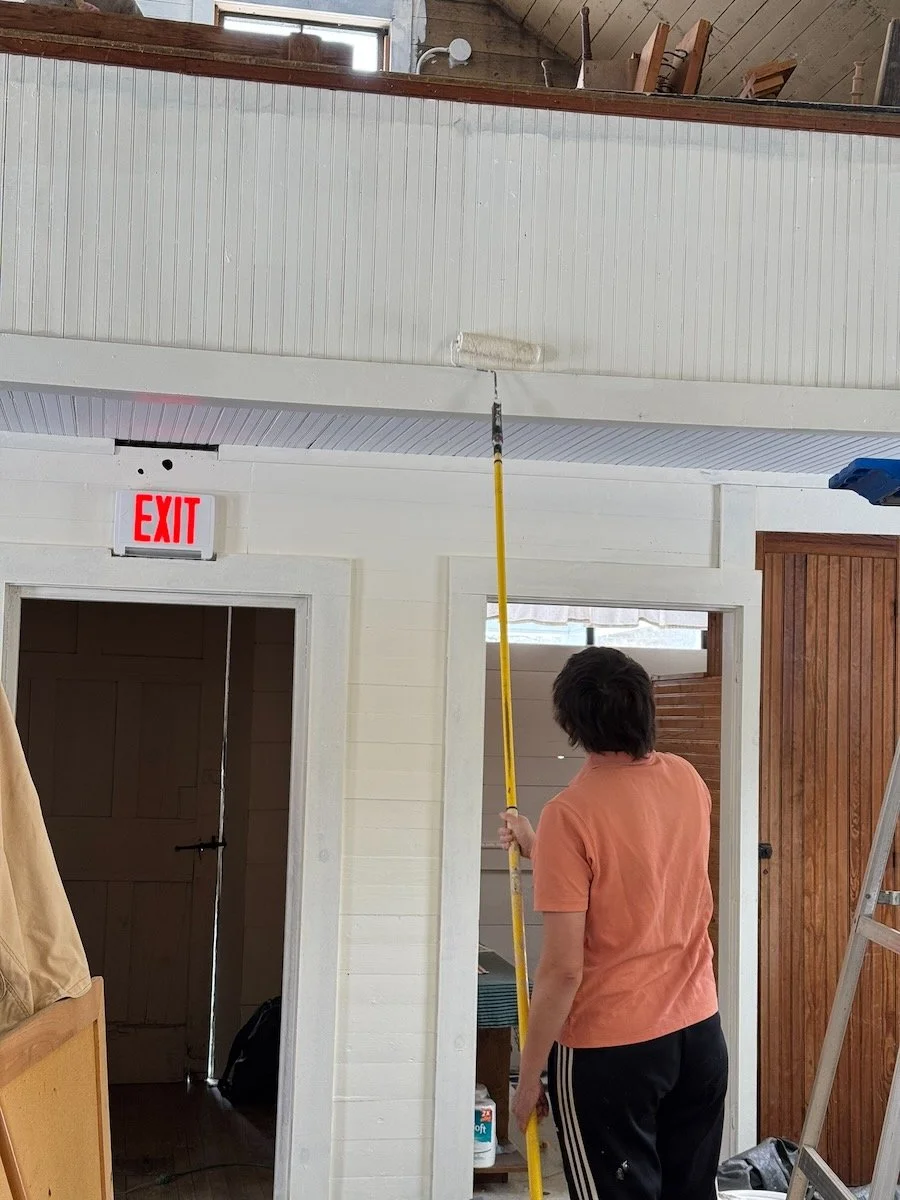 Person painting the ceiling with a paint roller using a long extension pole in a room with white walls, a ladder, and an exit sign.