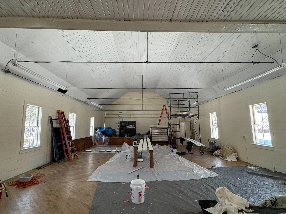 Interior of a large room under renovation with construction tools, materials, scaffolding, ladders, and plastic coverings on the floor.