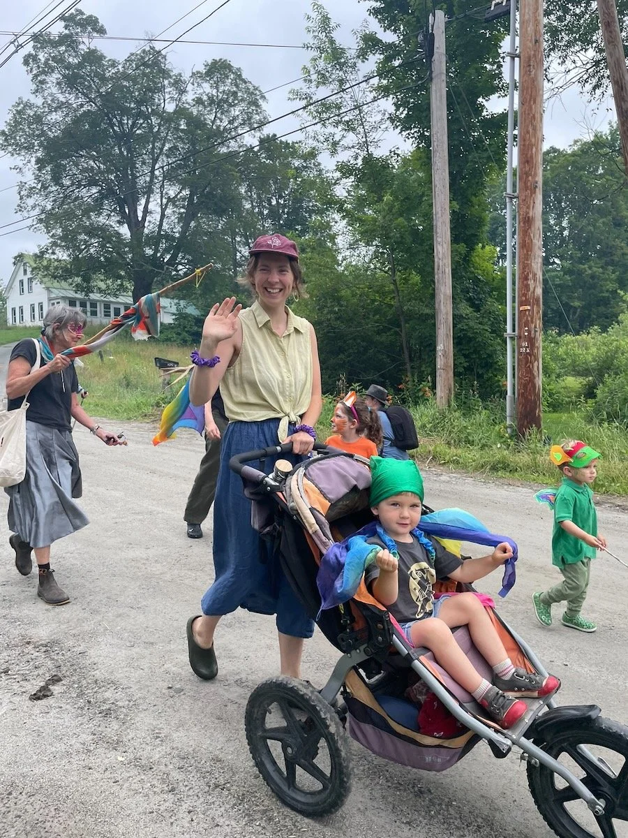 A woman smiling and waving as she pushes a stroller with a young girl dressed as a superhero during a parade or outdoor event, with children in costumes and face paint walking along a gravel road surrounded by trees and utility poles.