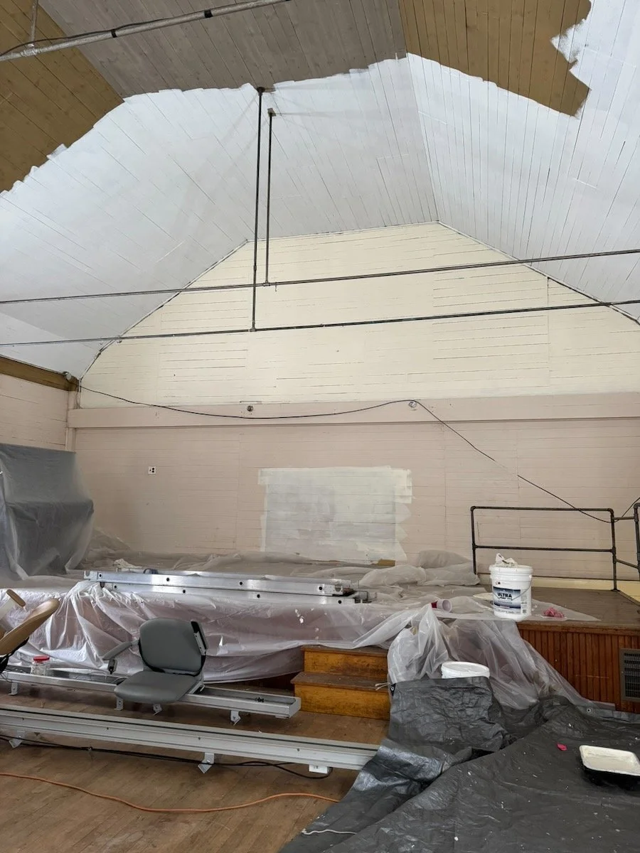Interior view shows a room under renovation with walls partially painted, covered with plastic sheeting, a ladder, paint buckets, and tools on the floor.
