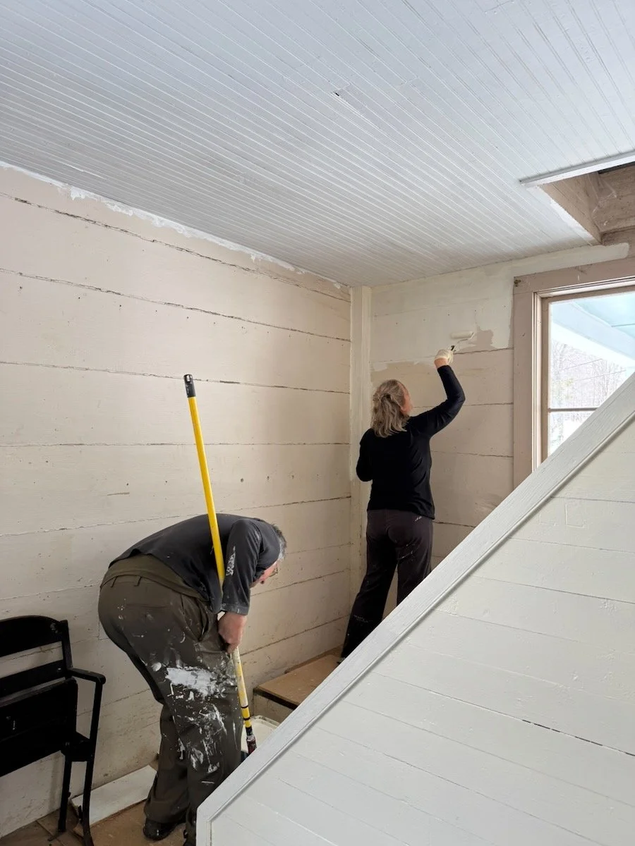 Two people working on the interior of a house. One is painting the wall near the window, the other is cleaning the floor with a broom. The room has unfinished walls and ceiling, with construction materials around.