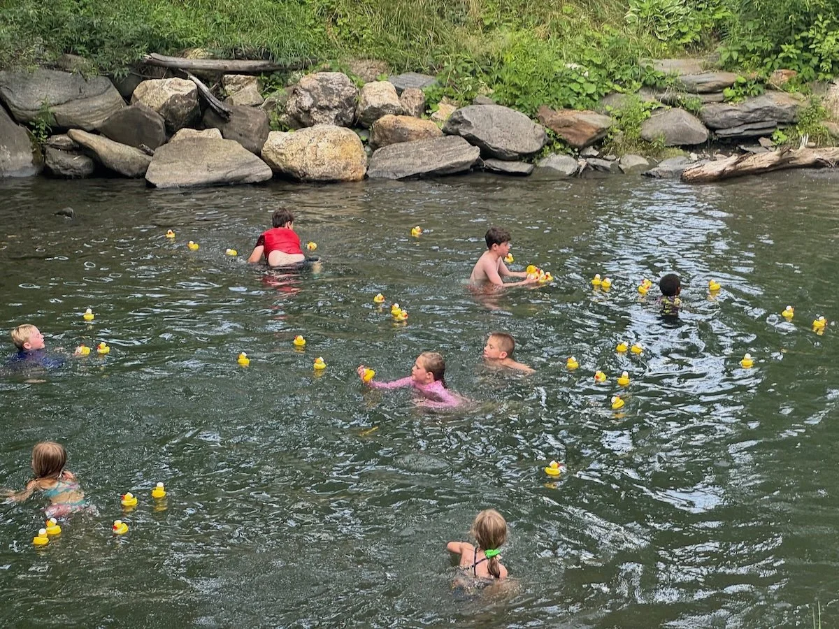 Children playing in a river, floating among yellow rubber ducks, with rocks and green foliage on the riverbank.