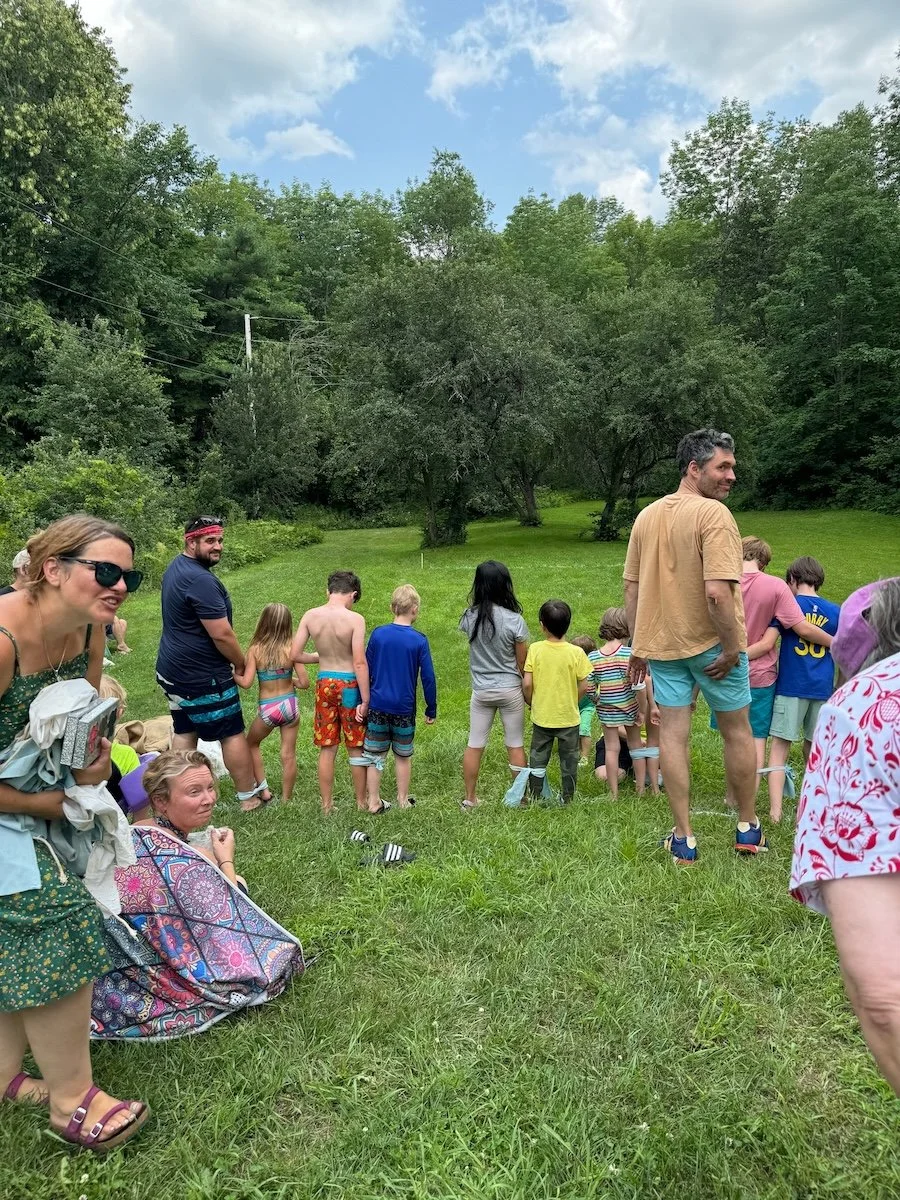 Group of children and adults outdoors on a grassy field, some in swimsuits and casual clothing, standing in a line, with lush green trees and partly cloudy sky in the background.