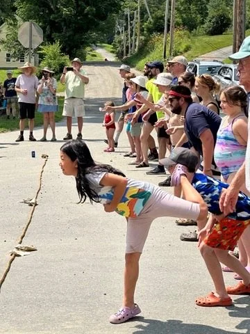 Children and adults playing a game of tug-of-war on a street during daytime, with trees and people watching.