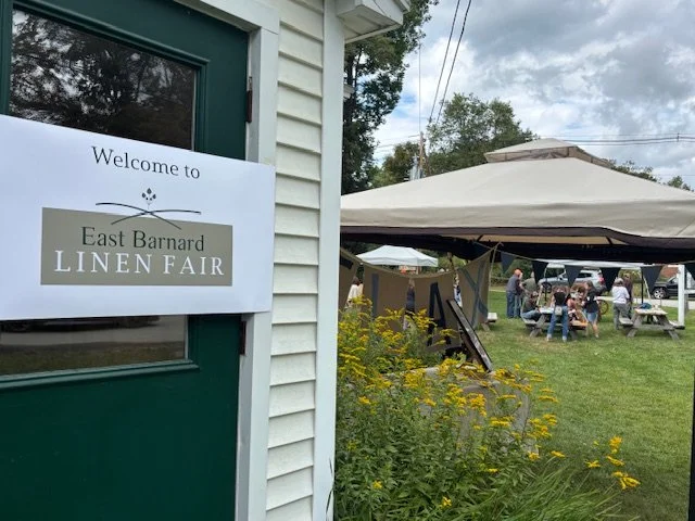 Sign on building reads 'Welcome to East Barnard Linen Fair,' with tents, flowers, and people gathering outdoors in a grassy area.