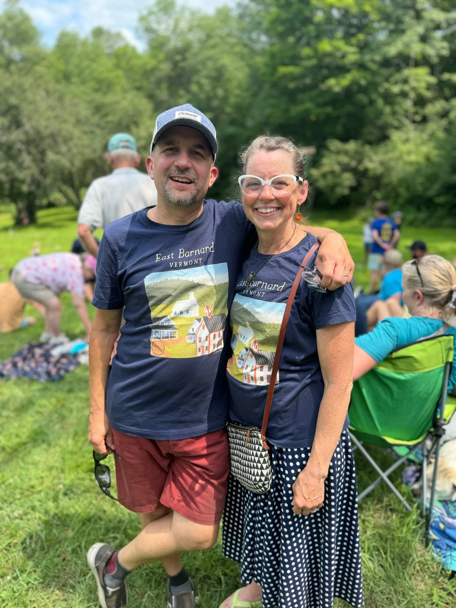 A smiling man and woman standing outdoors, embracing each other, with a group of people seated on the grass in the background during daytime. The man is wearing a baseball cap and a T-shirt that says 'East Barnard Vermont,' while the woman is wearing