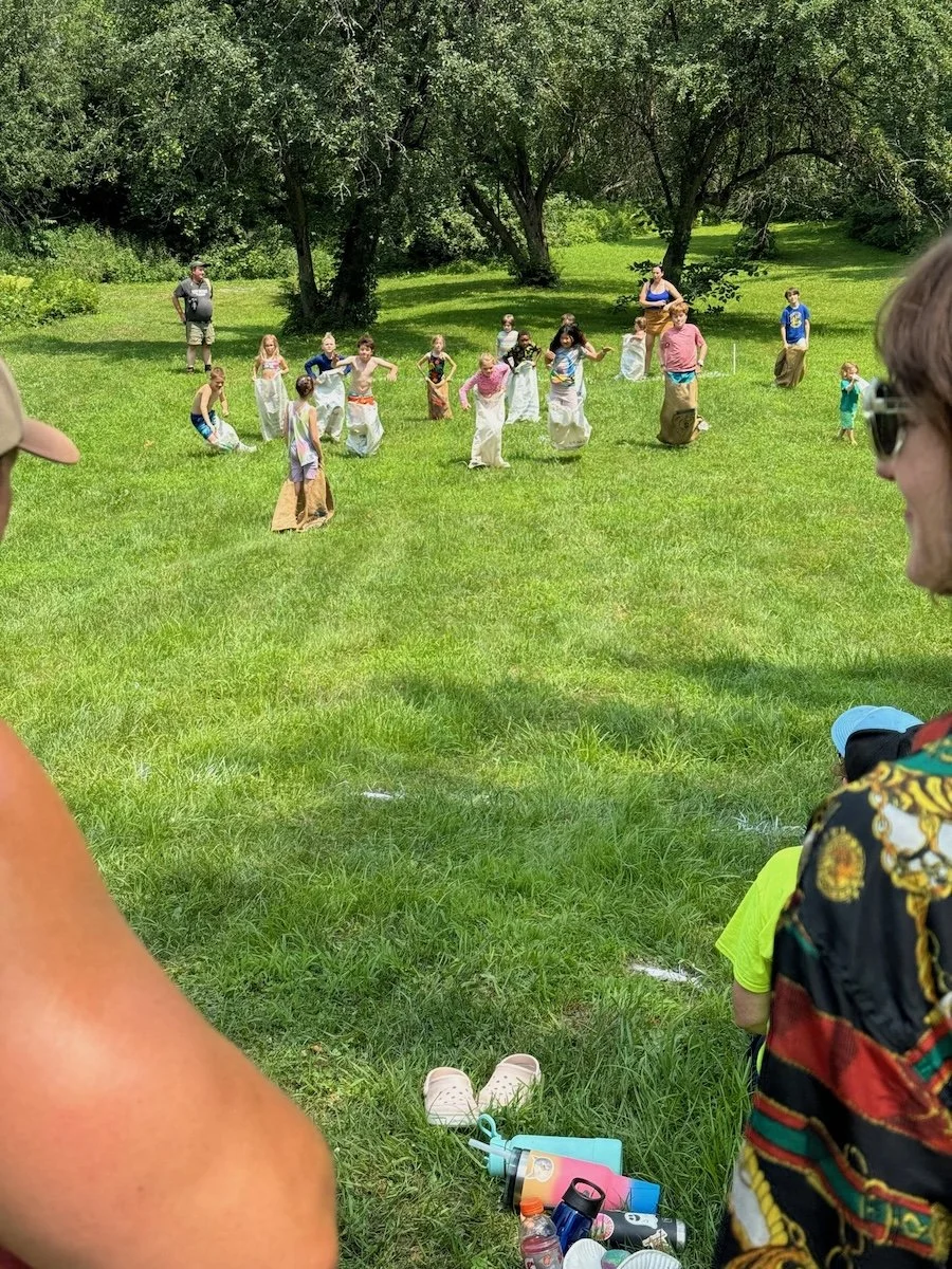 Children playing sack race outdoors on a sunny day in a grassy park with trees, with onlookers in the foreground.