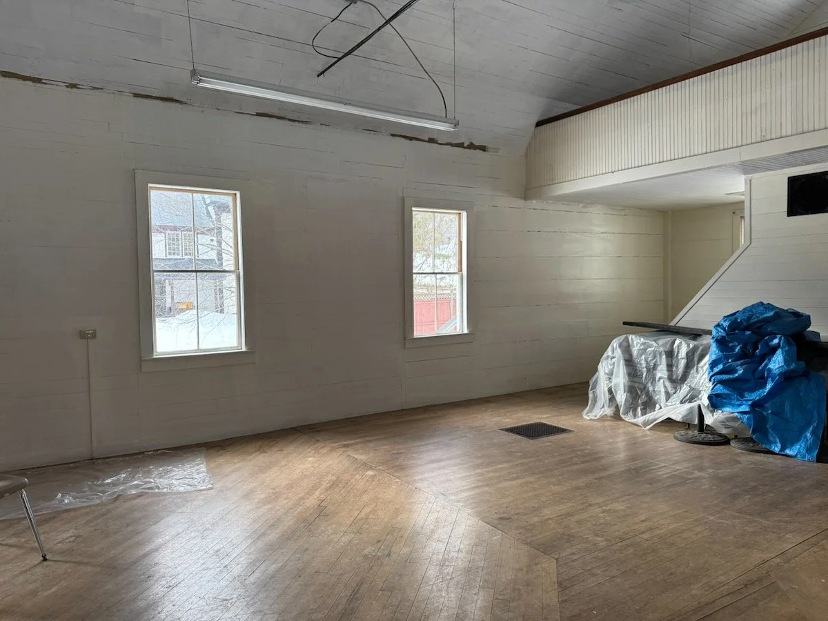 Empty room with two windows, a wooden floor, and a ceiling with hanging light fixtures. Some construction or renovation materials are covered with plastic and blue tarps.